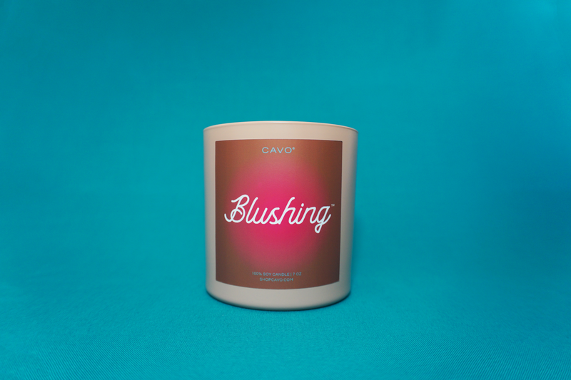 Blush pink jar with brown label with hit pink gradient circle reading "Blushing." by CAVO a Trendy Black Woman Owned, Black Owned Brand, Black Owned Candle Company. Candle sits on Aqua Blue backdrop.