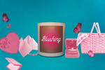 Blush pink jar with brown label with hit pink gradient circle reading "Blushing." by CAVO a Trendy Black Woman Owned, Black Owned Brand, Black Owned Candle Company. Candle sits on Aqua Blue backdrop. Candle is surrounded by collage of pink items including butterflies, chocolate box, mystery origami paper, BFF charm bracelet, blush compact, engagement ring, picnic basket